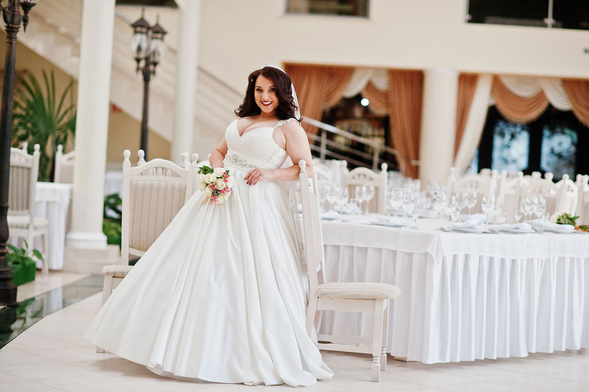 Vor Glück strahlende Braut in traumhaftem Hochzeitskleid steht in einem festlich vorbereitetem Saal und lehnt an einem Stuhl