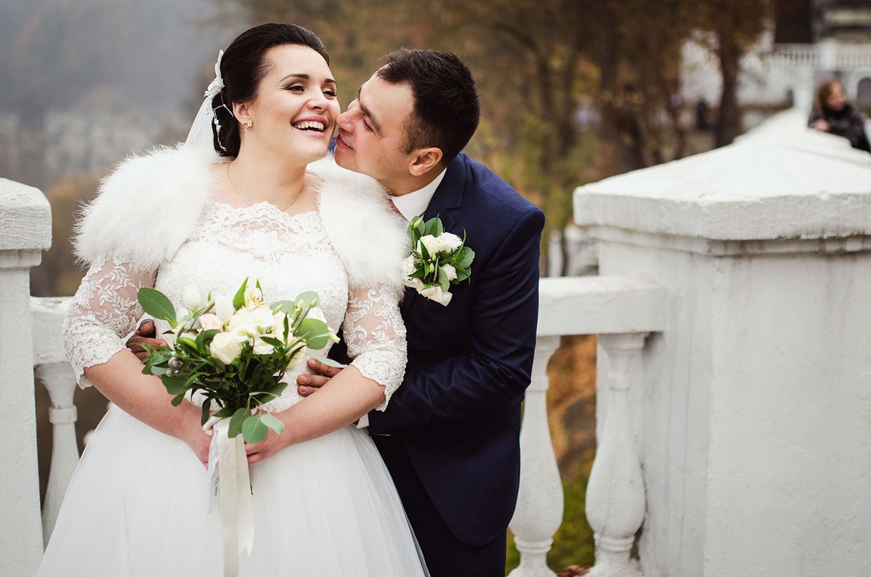 Braut mit Blumenstrauß lächelt glücklich, während ihr Ehemann sie umarmt und ihr einen Kuss auf die Wange gibt