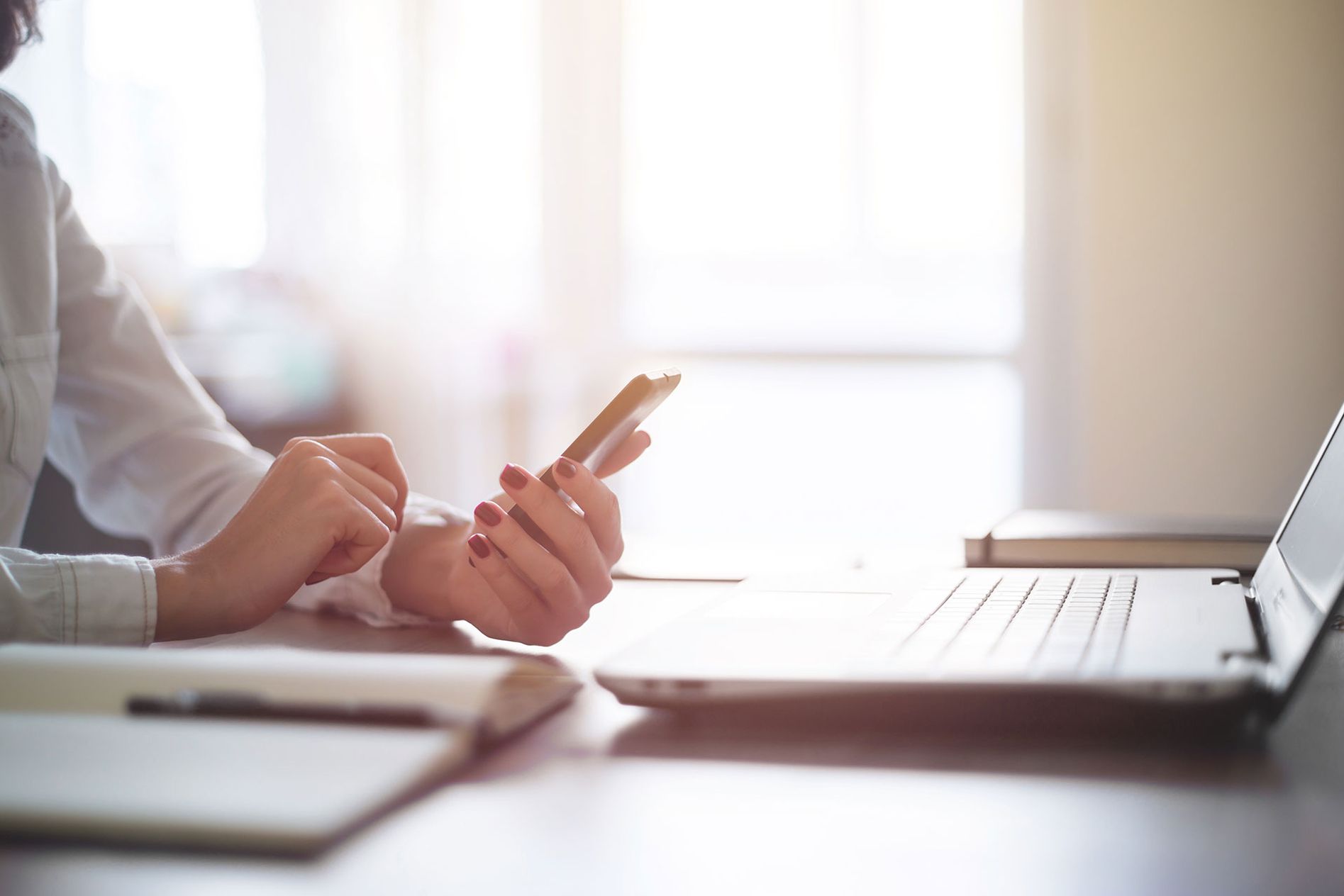 Eine Person hält ein Handy in der Hand, dahinter ist ein geöffneter Laptop zu sehen